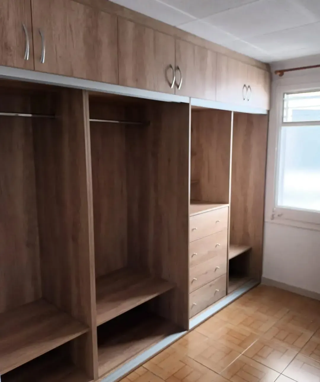 Armario de madera con puertas correderas y estantes, junto a una ventana en una habitación con suelo de baldosas.