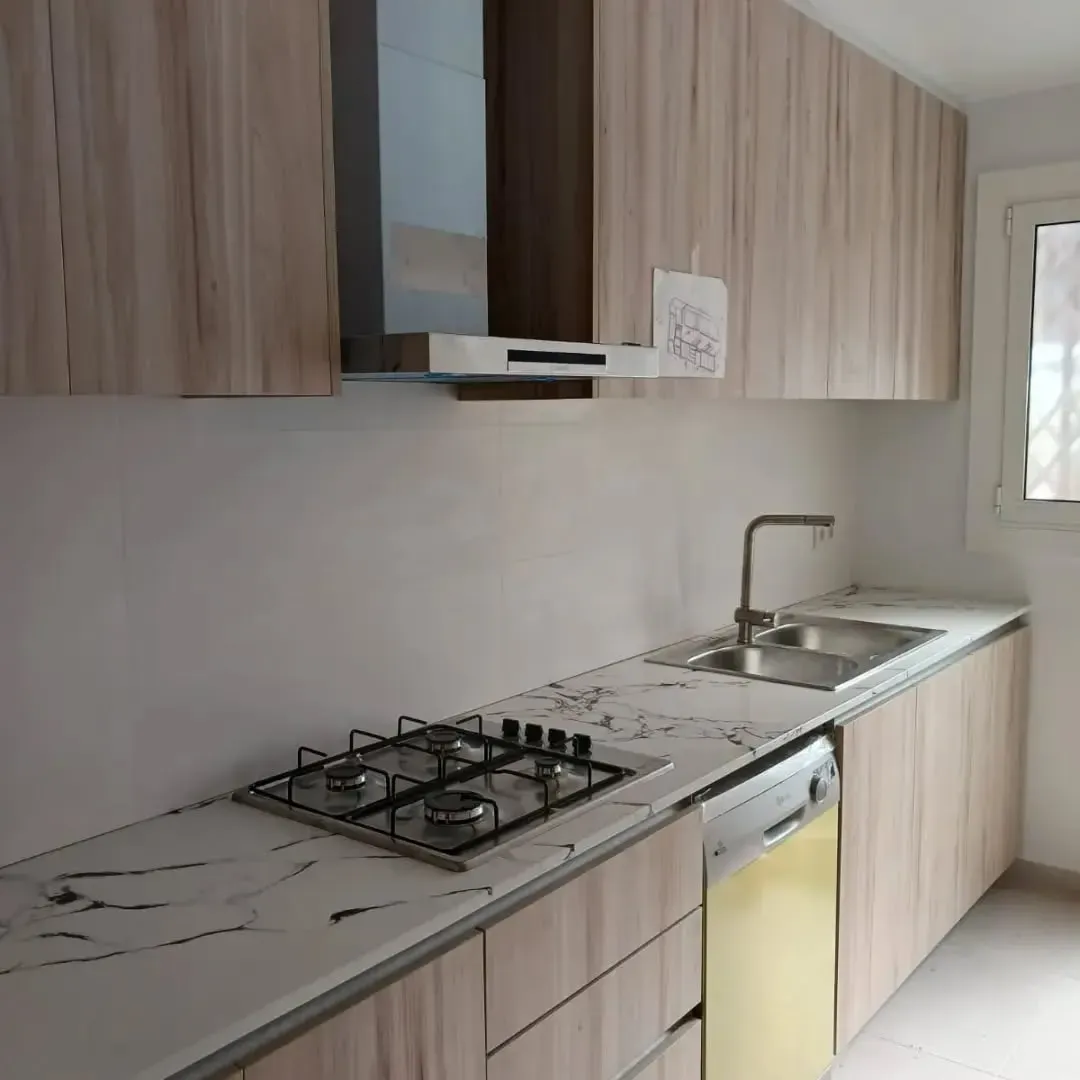 Cocina con gabinetes de madera clara, encimera blanca con vetas grises, estufa a gas y fregadero.