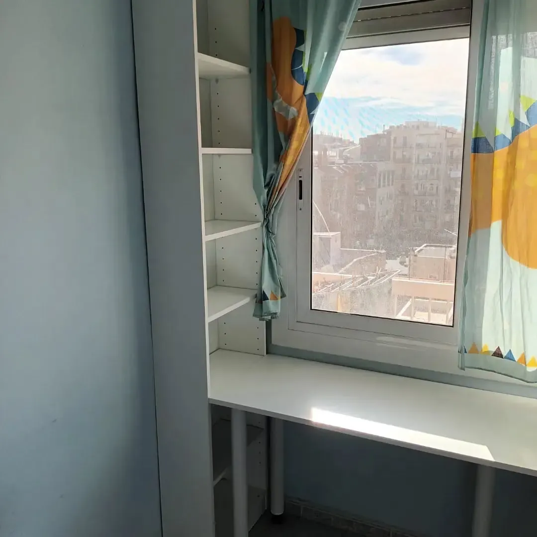 Un escritorio blanco y una estantería junto a una ventana con cortinas azules, con vistas a una ciudad.