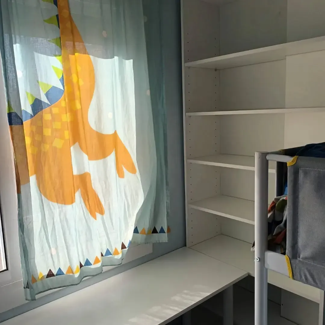 Estantería blanca junto a una litera. A la izquierda, una ventana con una cortina de dinosaurios.