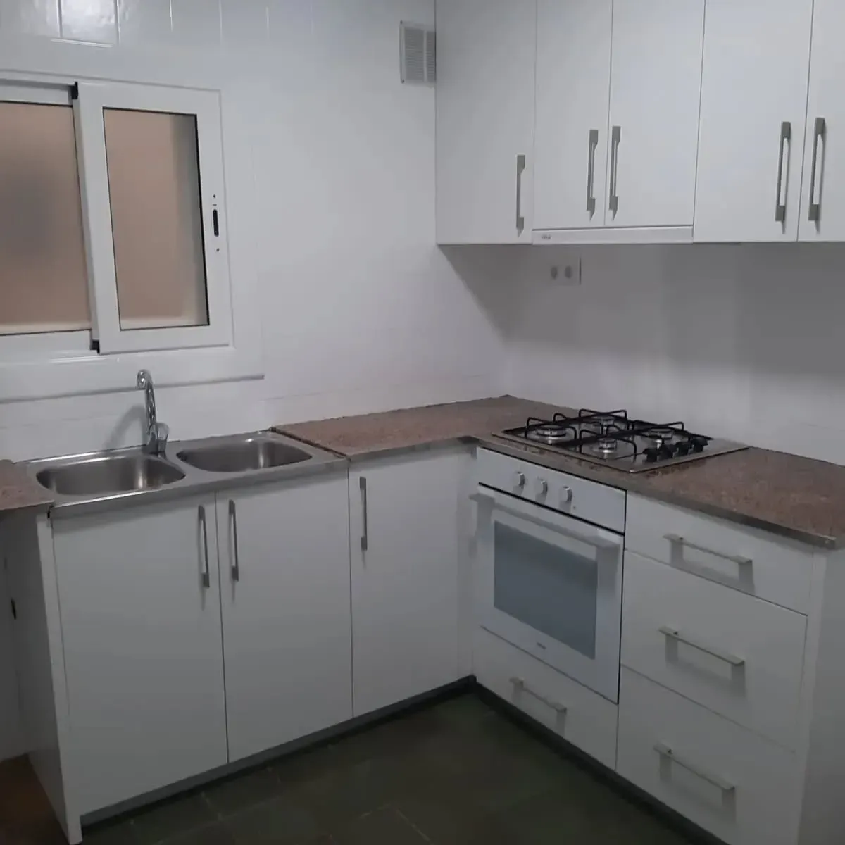 Muebles de cocina blancos, encimera, fregadero y horno. Una ventana a la izquierda.