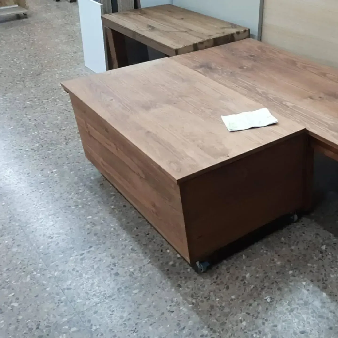 Escritorio de madera marrón con armario de almacenamiento adjunto y ruedas sobre un piso moteado.