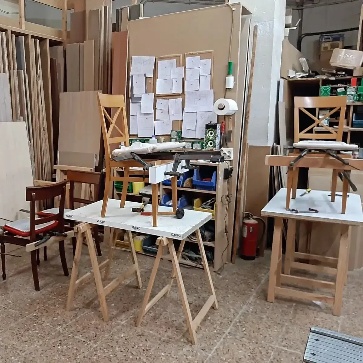 Escena de taller con sillas, caballetes y herramientas; láminas de madera y notas en la pared.