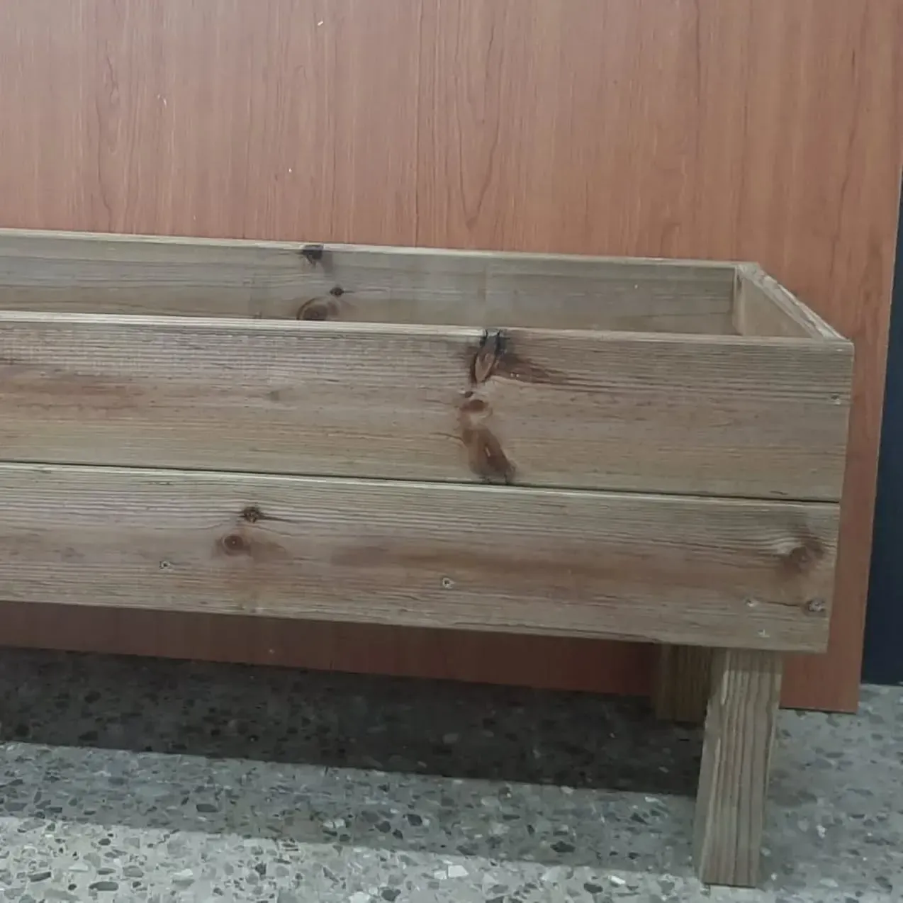 Jardinera de madera con patas, de color marrón, sobre pared marrón.