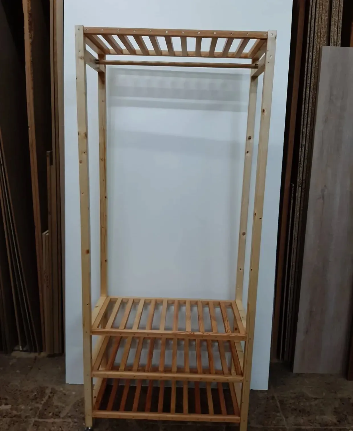 Perchero de madera con barra para colgar y dos estantes, contra una pared blanca.