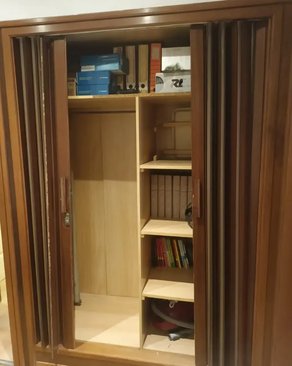 Armario de madera con puertas de acordeón abiertas, que revela estantes con libros, archivos y una aspiradora.