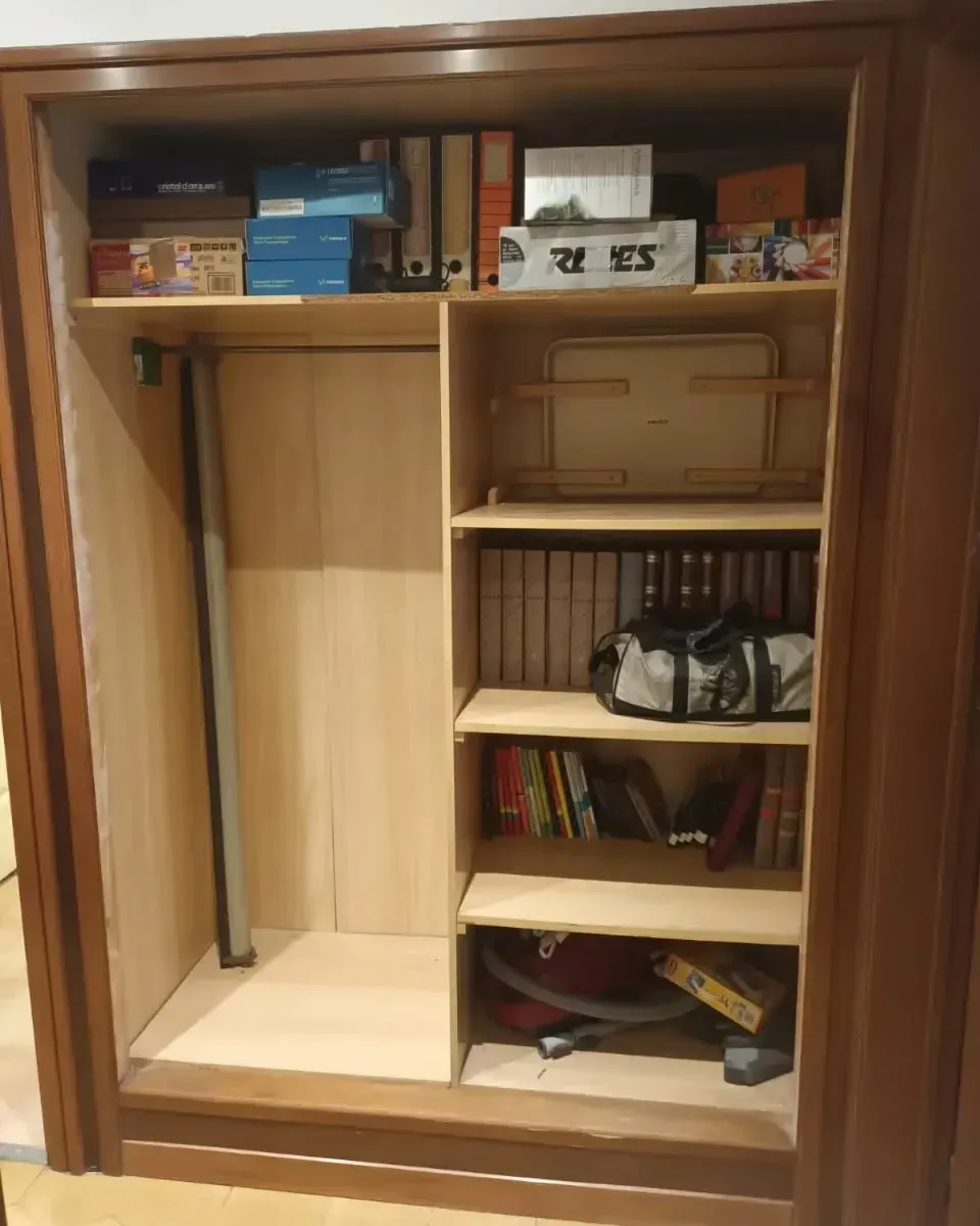 Armario de madera con estantes que contienen cajas, libros y una bolsa. En el estante inferior hay una aspiradora.