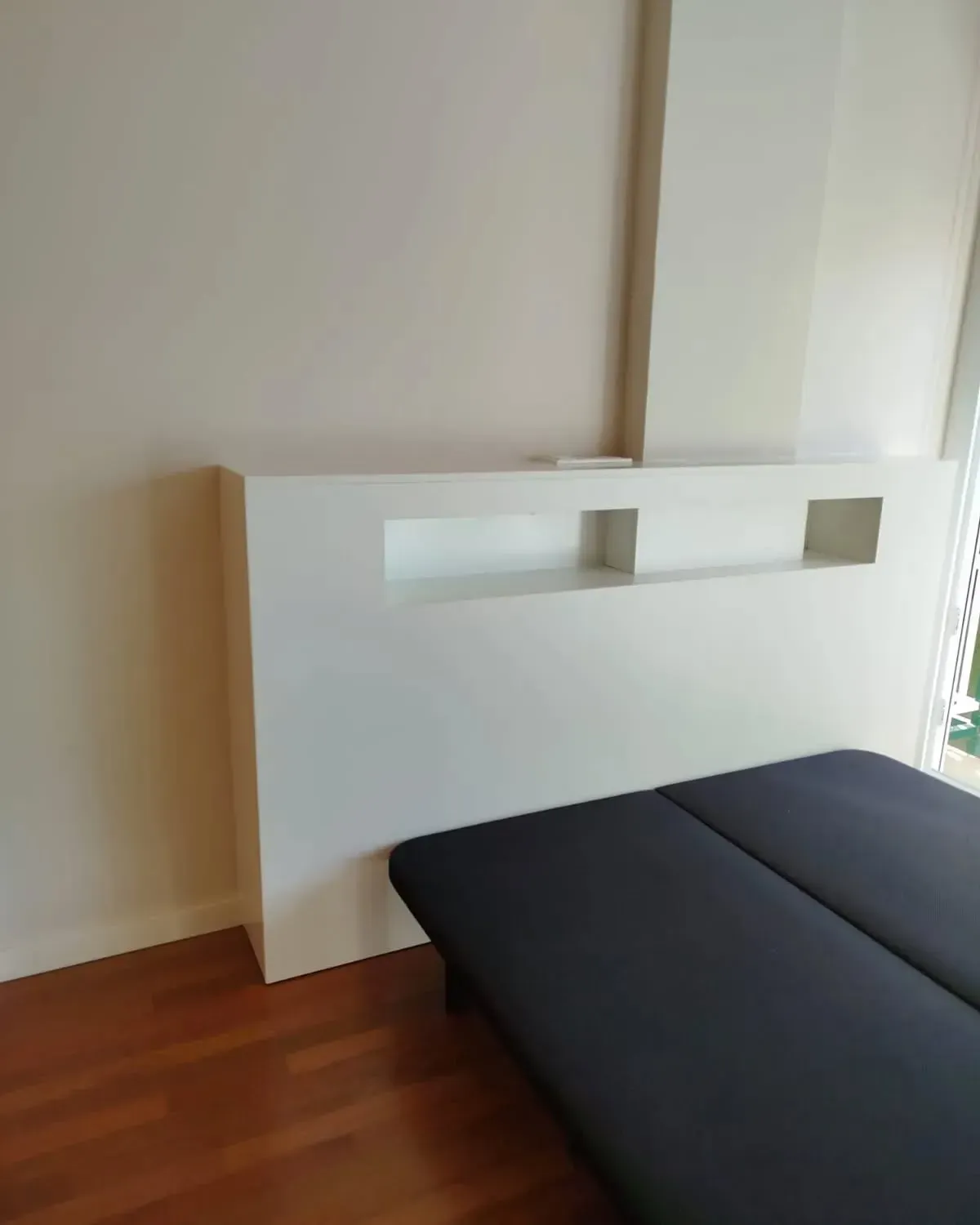 Cabecero blanco con estantes empotrados sobre una cama negra. Suelo de madera marrón.