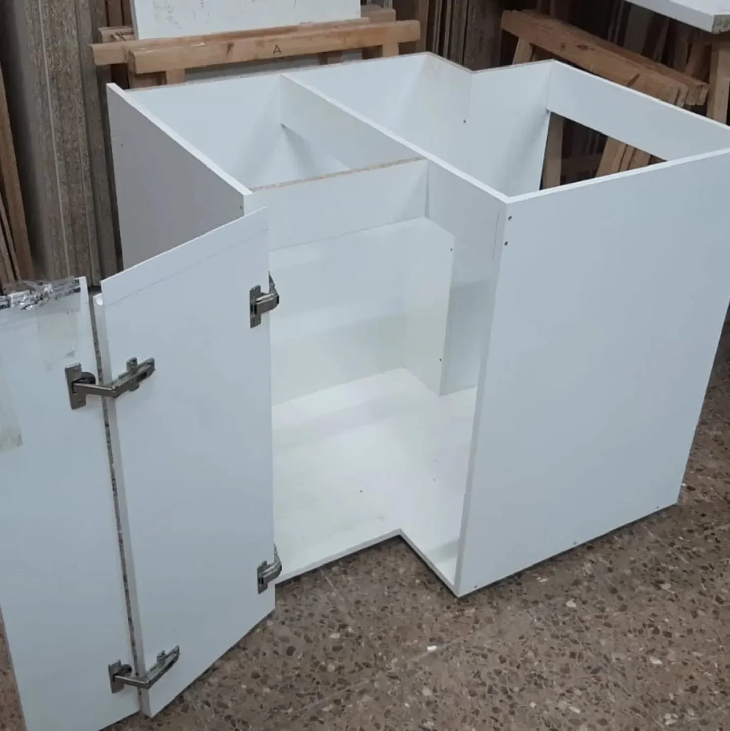 Mueble esquinero blanco, sin terminar, con puerta abierta y estantes interiores visibles.