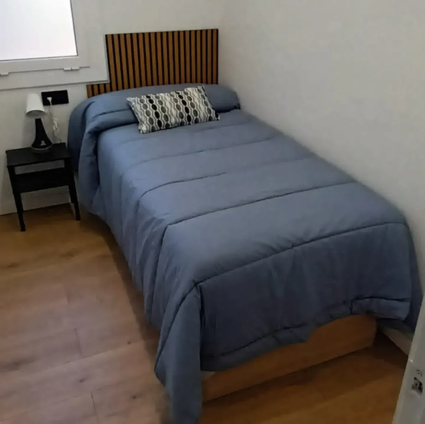 Un dormitorio pequeño con una cama individual cubierta con ropa de cama azul, cabecera de madera y mesita de noche negra.