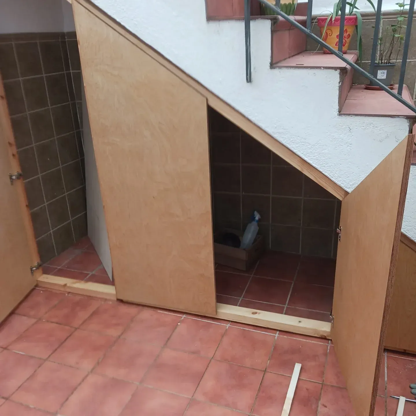 Trastero de madera construido bajo escalera exterior con suelo y paredes de baldosas marrones.