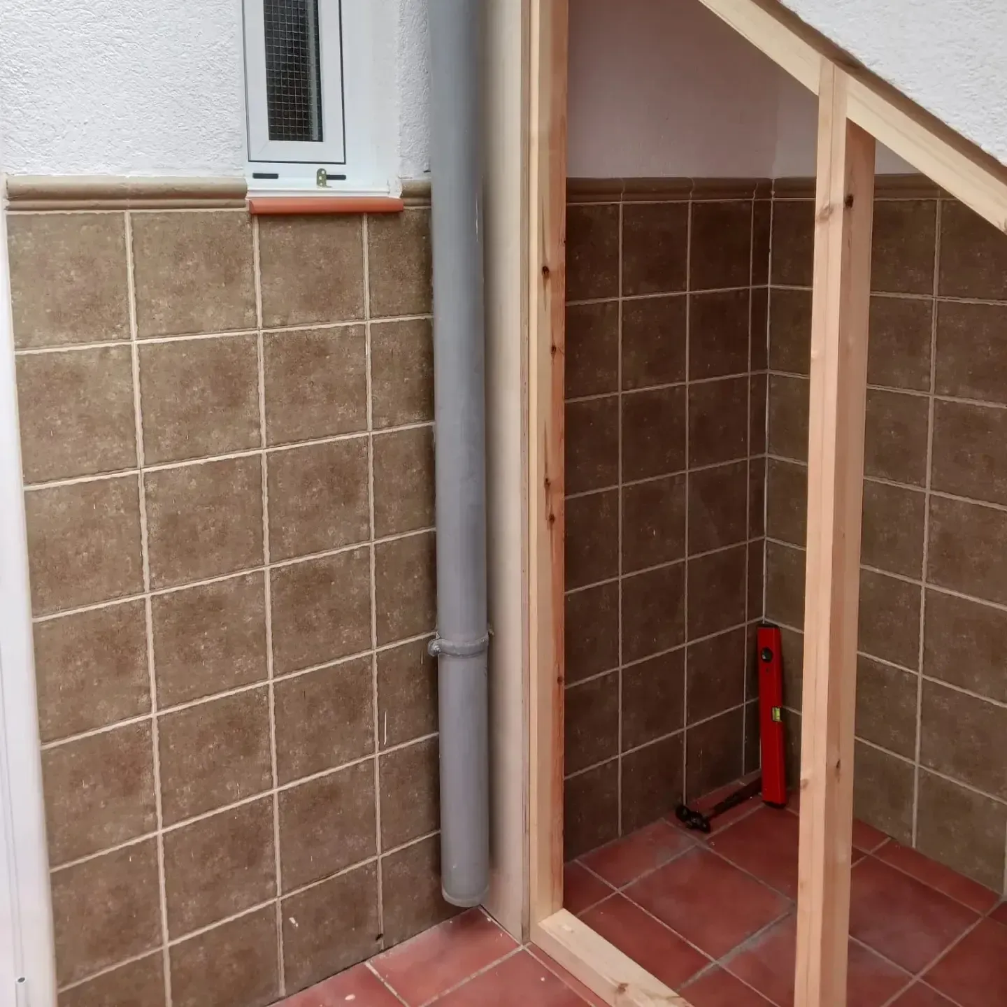 Renovación del baño en proceso. Paredes de azulejos marrones, tubería gris, estructura de madera y nivelado.