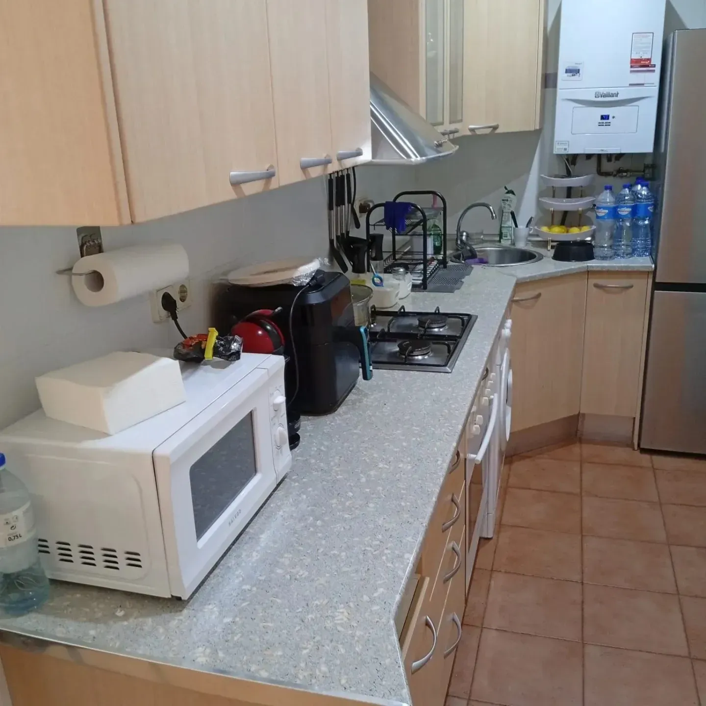 Cocina con gabinetes, encimera y electrodomésticos en tonos claros. Incluye microondas, estufa y calentador de agua.