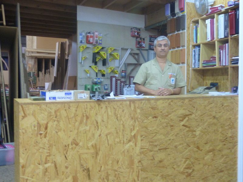 Hombre de pie detrás de un mostrador de madera en una ferretería; herramientas y estantes de suministros en el fondo.