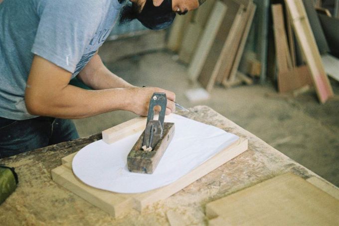 Carpintero que utiliza un cepillo de mano para dar forma a la madera; taller con madera al fondo.