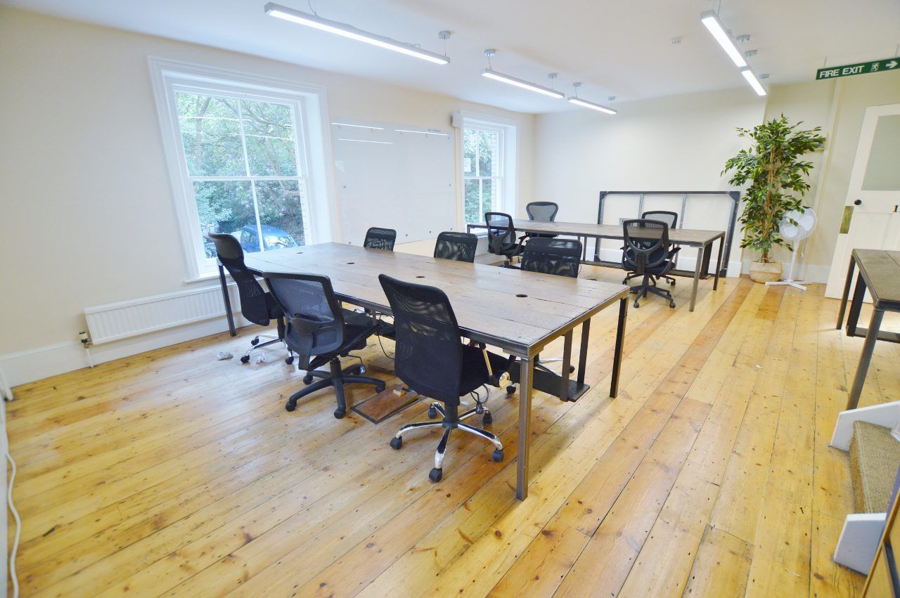 An office space with wooden floors, multiple desks, and black office chairs. Bright with natural light.