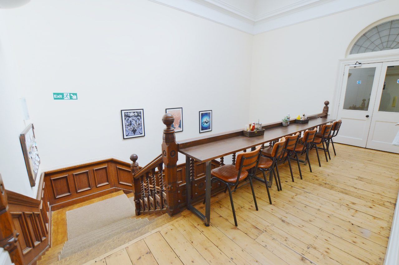 Wooden staircase next to a long table with chairs; white walls, posters, and a door.