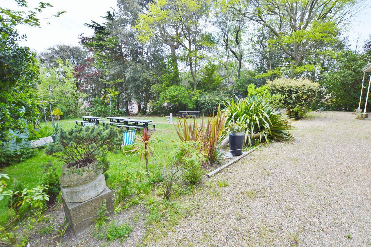 Lush green garden with gravel path, picnic tables, and various plants and trees under a cloudy sky.