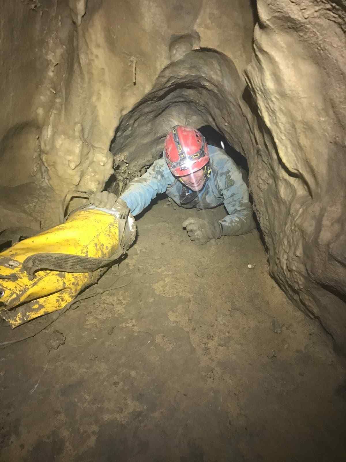 Espeleólogo con casco rojo arrastrándose por un estrecho túnel fangoso, tirando de una bolsa amarilla tras él.