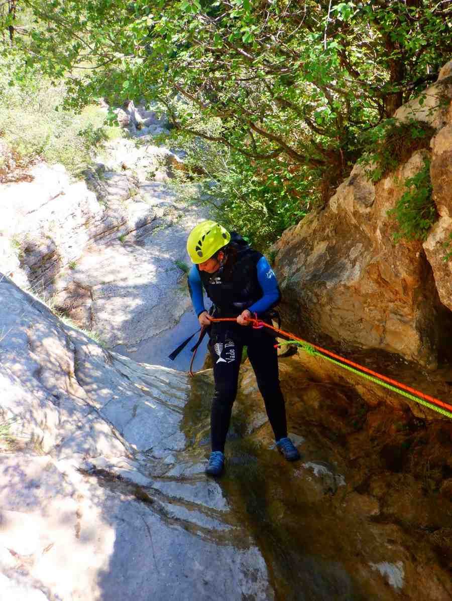 Un practicante de rápel con casco desciende por un estrecho cañón rocoso junto a un arroyo y árboles.