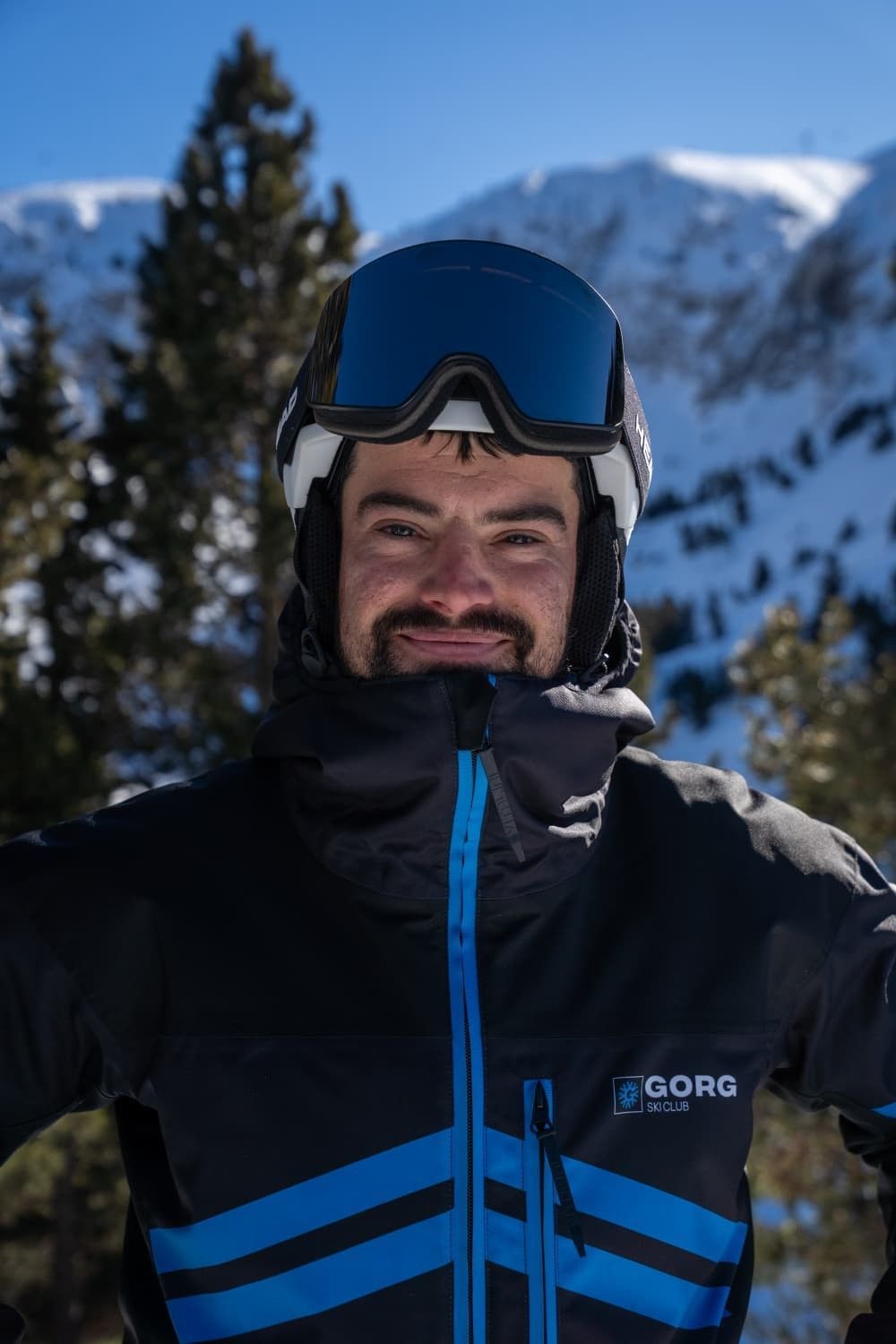 Esquiador sonriente con chaqueta negra y gafas, con montañas nevadas al fondo.