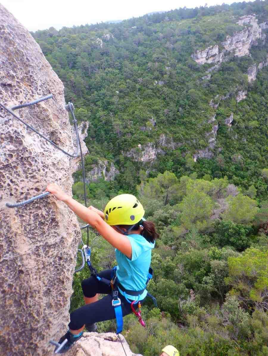 Escalador con camisa azul y casco amarillo ascendiendo por la pared de un acantilado sobre un valle verde.