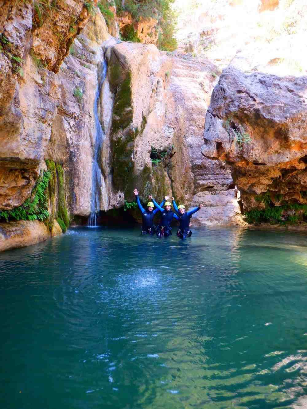 Personas con cascos azules flotando en una poza turquesa bajo una cascada rocosa.
