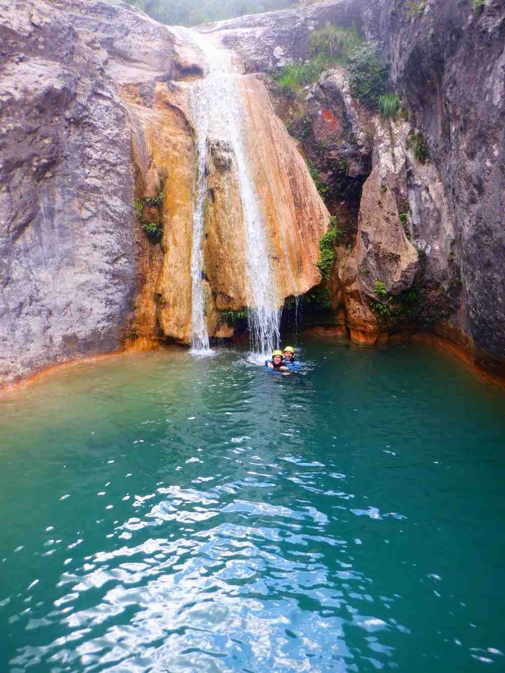 Una pequeña cascada cae en una poza de color turquesa entre acantilados rocosos, con un nadador cerca de la base.