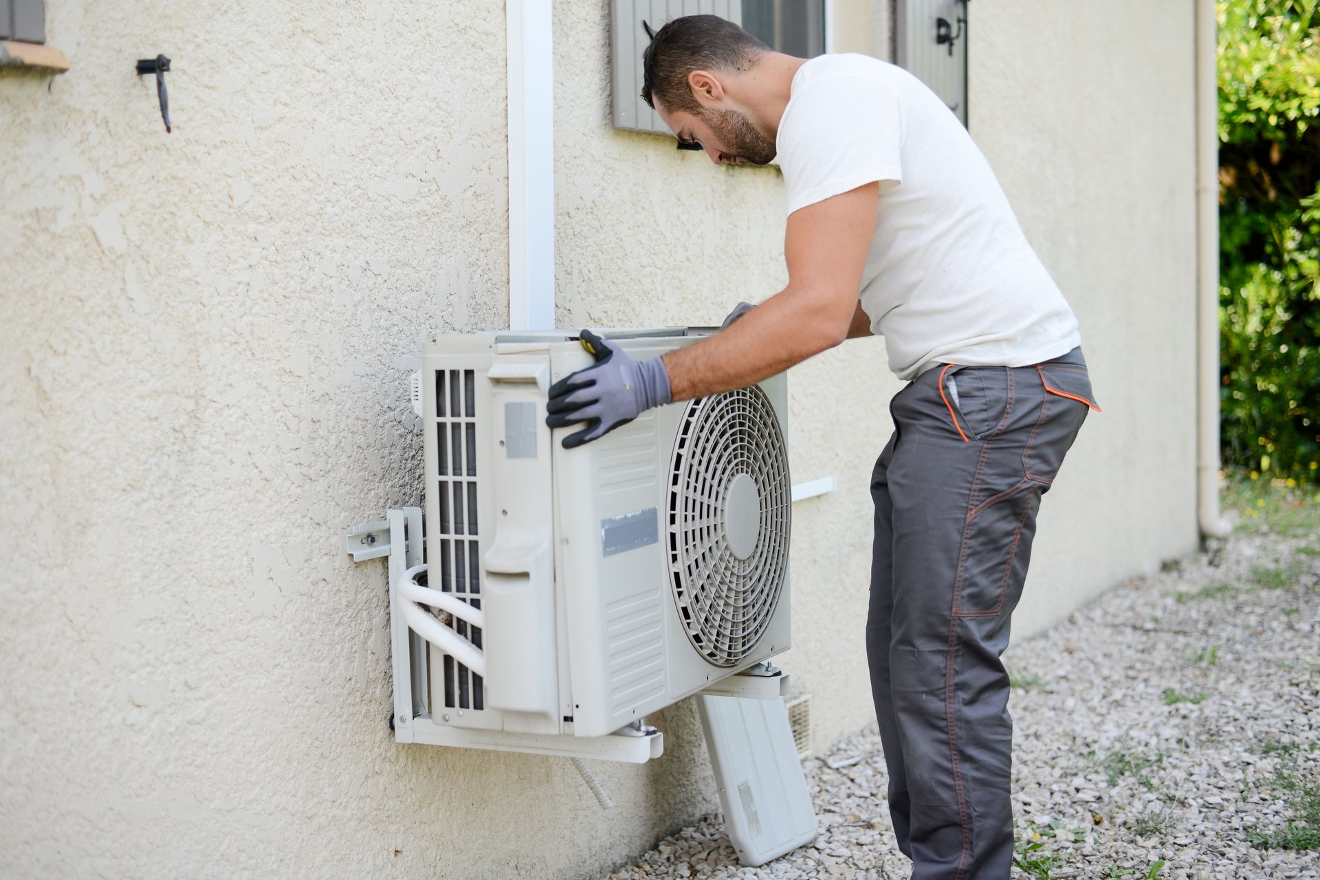 Un homme en pantalon de travail gris et chemise blanche installe un climatiseur à l'extérieur.