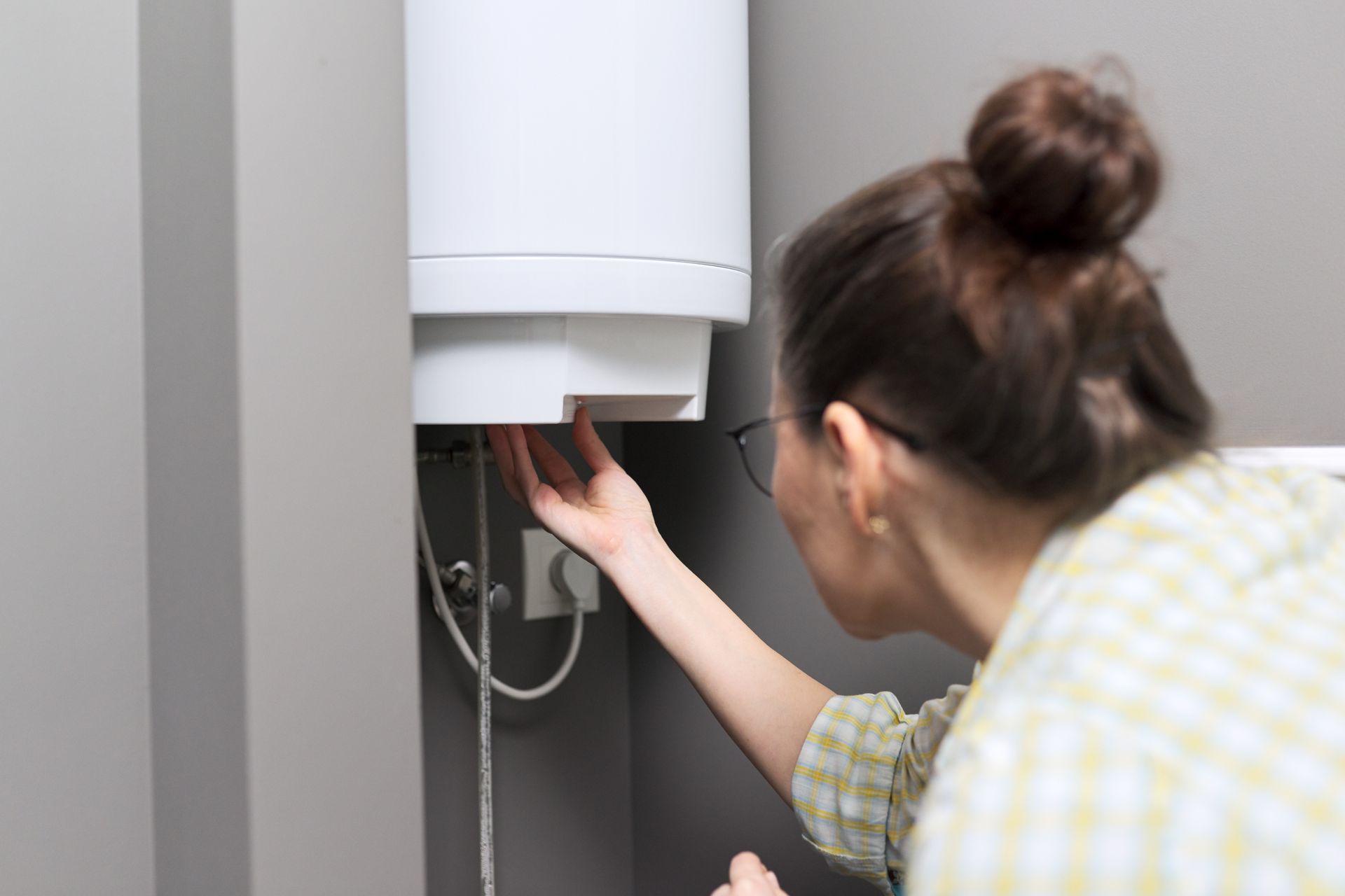 Une femme inspecte un chauffe-eau blanc, tendant la main vers le bas dans un espace aux murs gris.