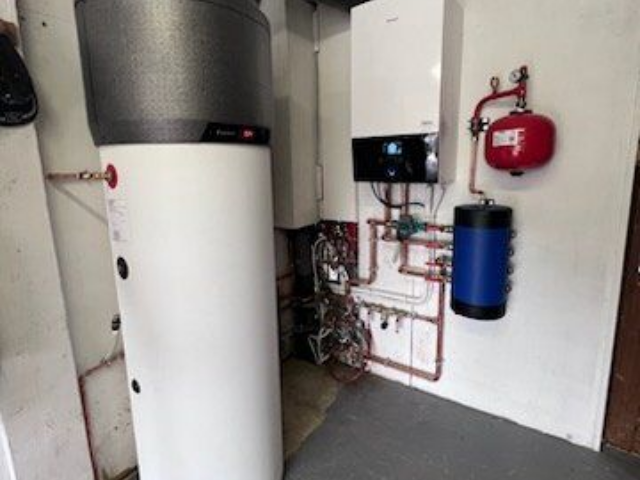 Chauffe-eau, chaudière et réservoirs blancs dans un local technique avec des tuyaux en cuivre et un vase d'expansion rouge.