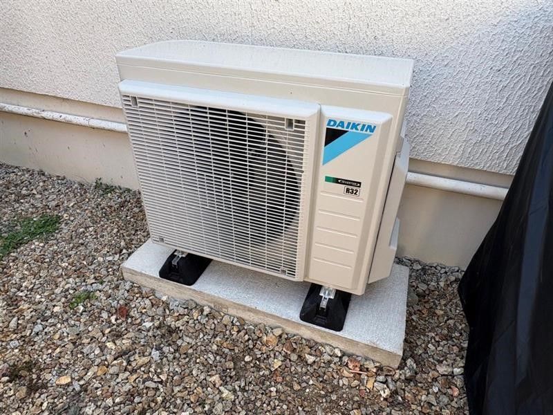 Pompe à chaleur Daikin fixée sur un socle en béton contre un mur clair. Sol en gravier.
