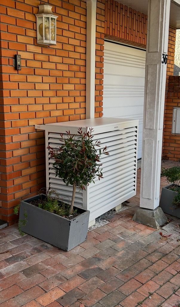 Enclos à claire-voie blanche abritant un objet, à côté d'un mur de briques rouges et d'une plante en pot sur une terrasse en briques.