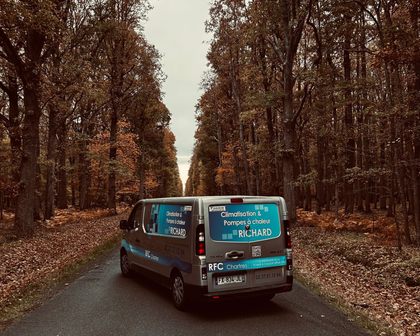 Une camionnette roule sur une route bordée d'arbres en automne. La camionnette est argentée et les arbres ont des feuilles brunes et orangées.