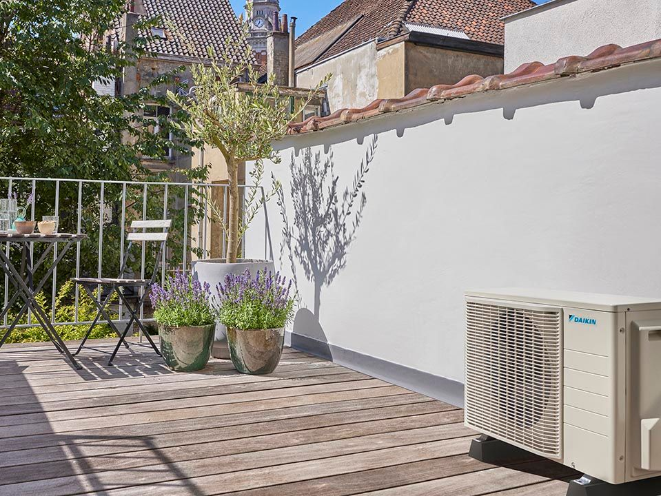 Terrasse avec mobilier de jardin, plantes en pot et unité de climatisation adossée à un mur blanc.