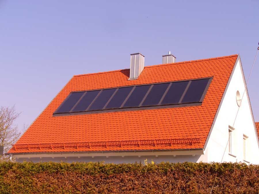 Zwei Arbeiter in orangefarbenen Sicherheitswesten und Schutzhelmen installieren Solarpaneele auf einem Dach.