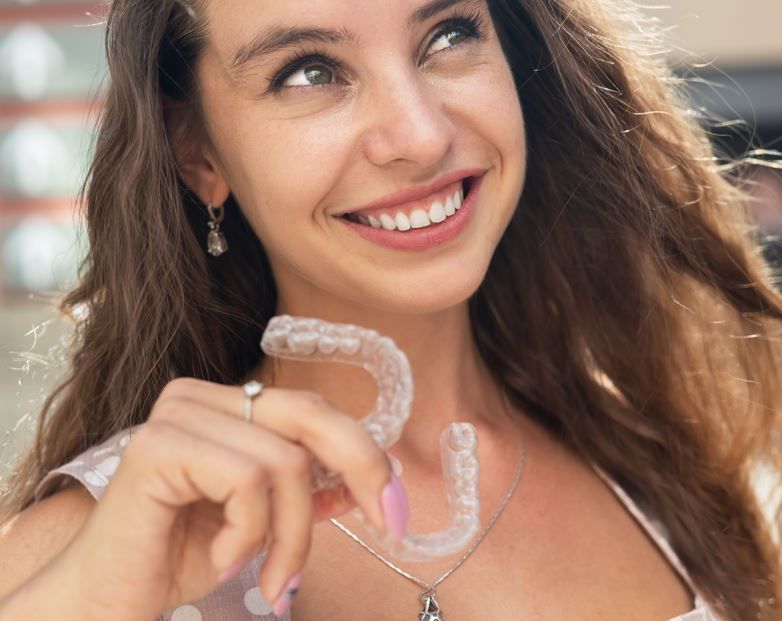 Mujer sonriente sosteniendo alineadores dentales transparentes al aire libre, con un suave fondo nevado.