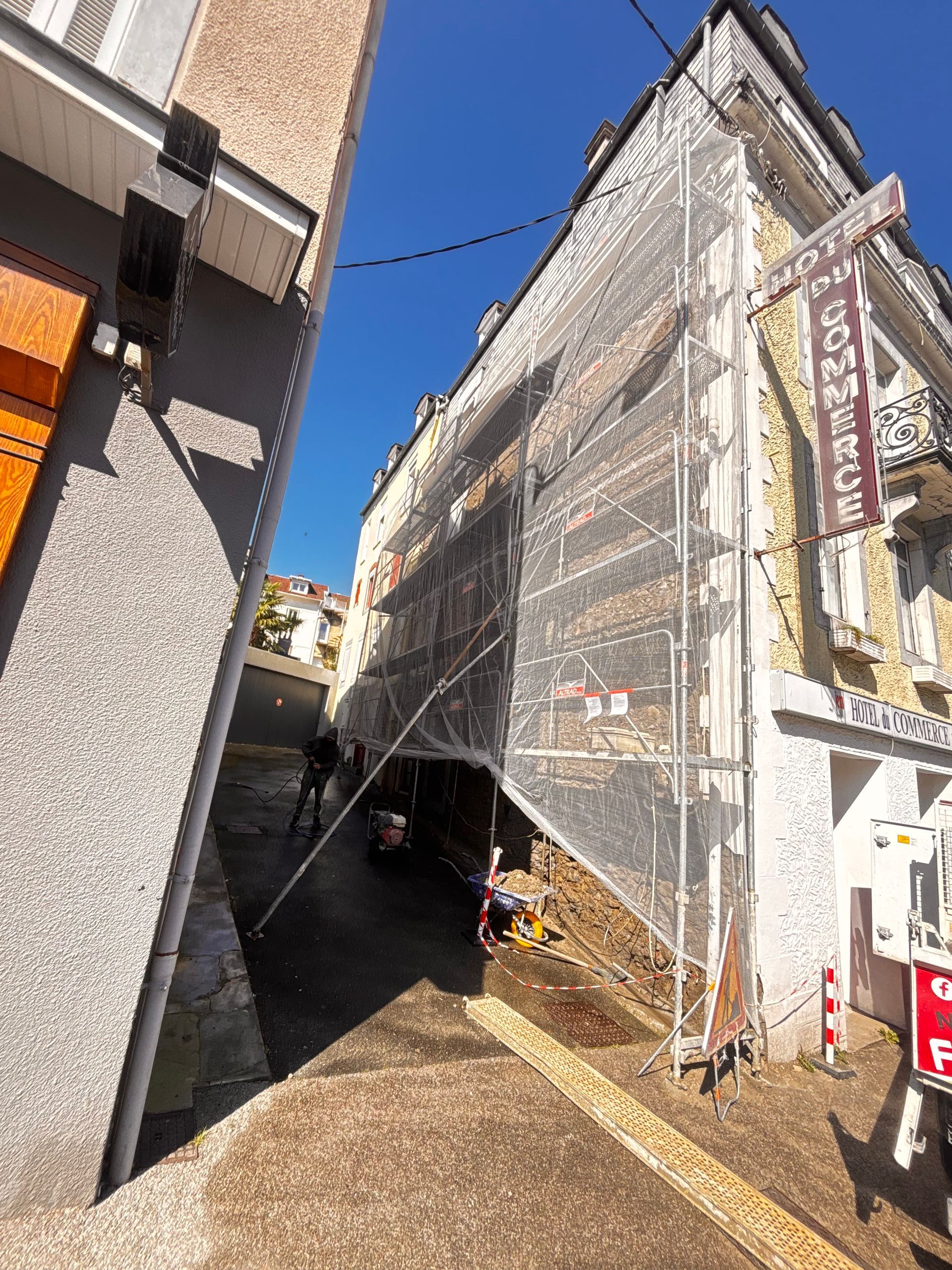 La façade d'un immeuble recouverte d'échafaudages et de filets de protection, à côté d'une ruelle étroite sous un ciel d'un bleu éclatant.