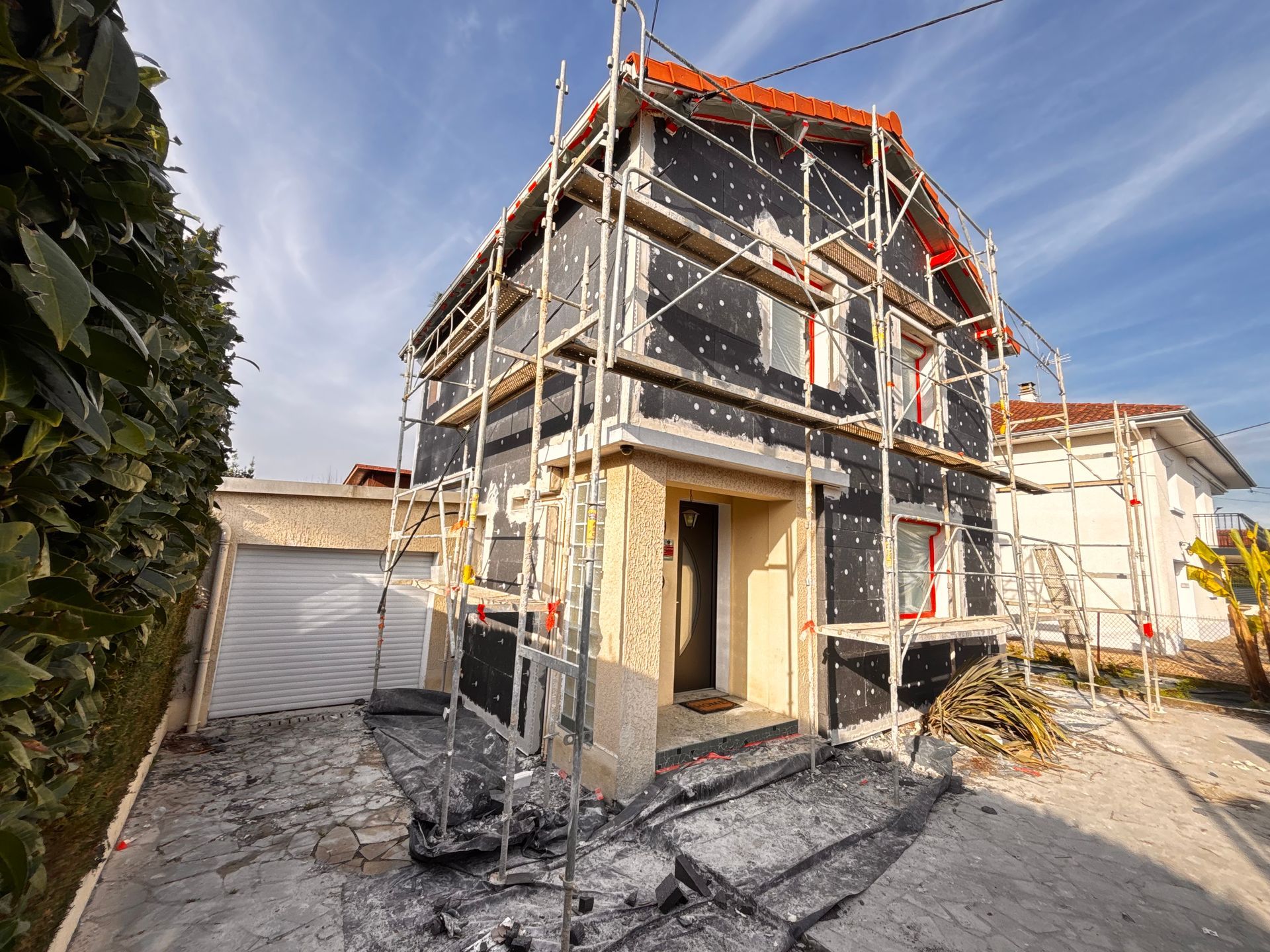 Une maison en rénovation, recouverte de panneaux isolants sombres et entourée d'échafaudages, par une journée ensoleillée.