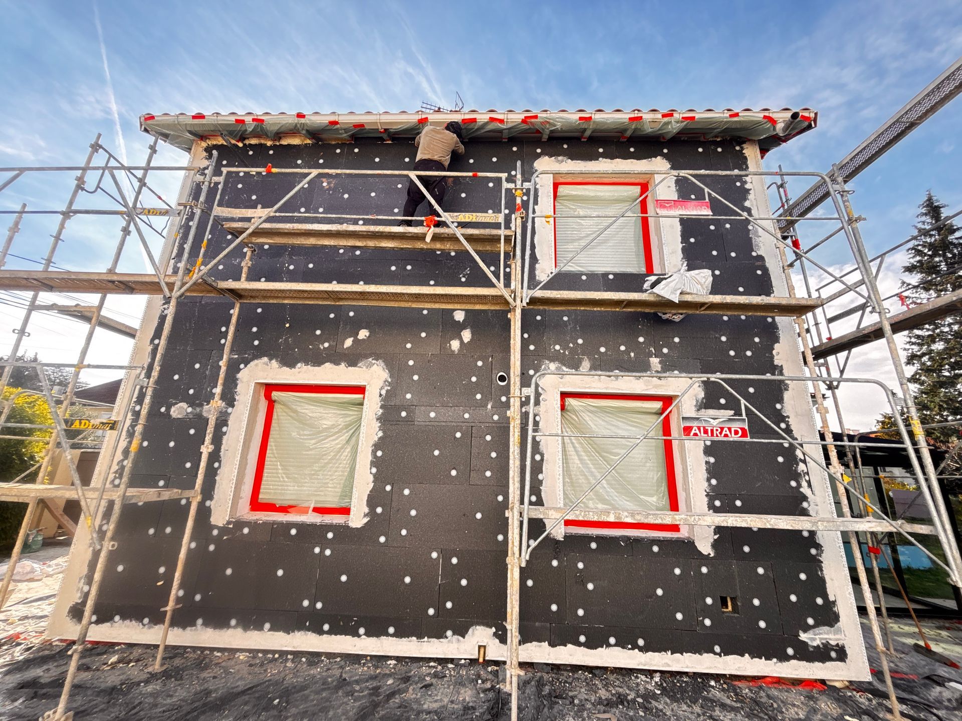 Un ouvrier, juché sur un échafaudage, installe des panneaux d'isolation noirs sur le mur extérieur d'une maison à deux étages.