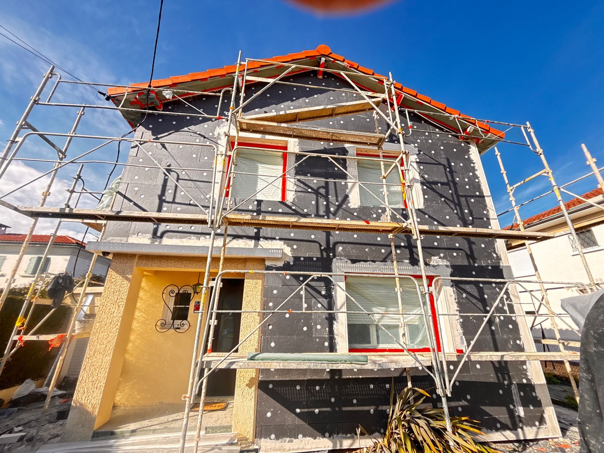L'extérieur d'une maison est recouvert de panneaux isolants noirs, encadré par une structure métallique d'échafaudages, sous un ciel d'un bleu éclatant.