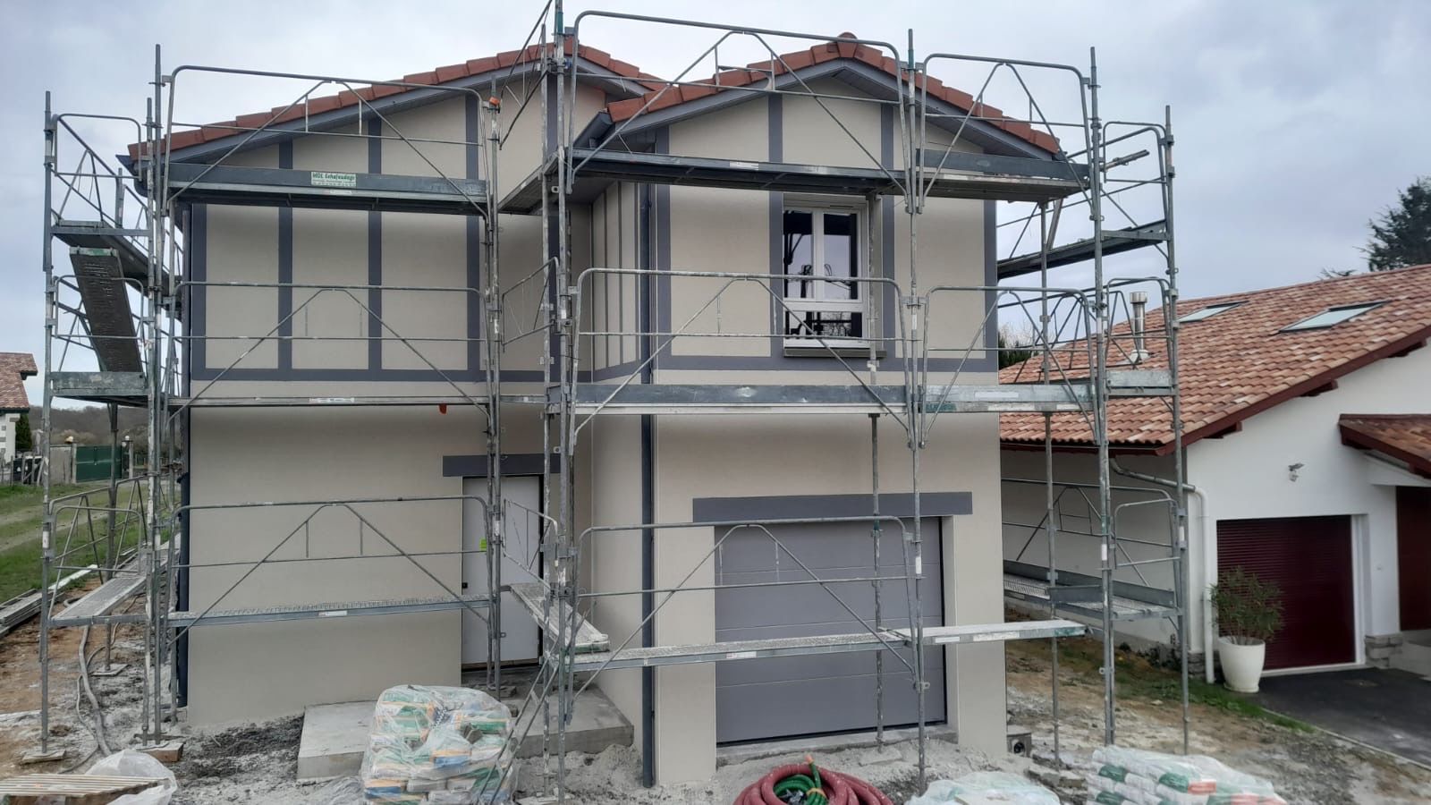 Une maison à deux étages en construction, recouverte d'échafaudages, avec des murs en stuc beige et des boiseries gris foncé.