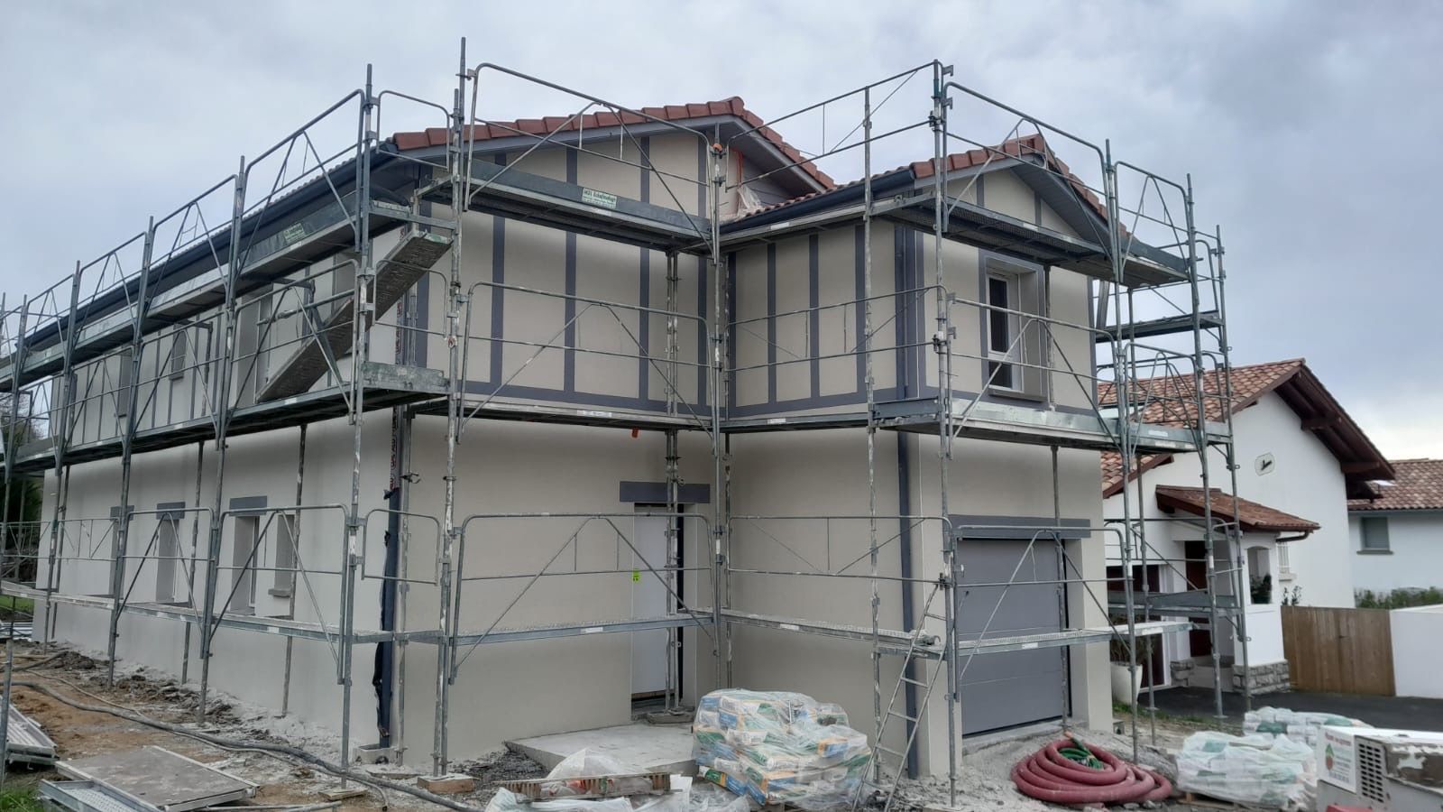 Une maison à deux étages en construction, enveloppée d'échafaudages métalliques, avec des murs extérieurs de couleur claire et un toit de tuiles.
