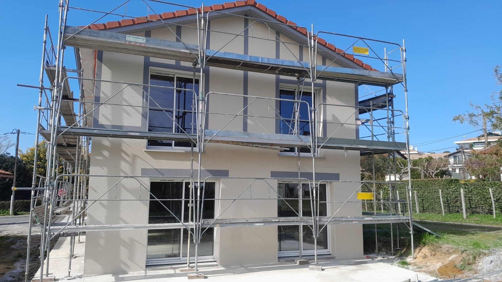 Une maison de banlieue beige à deux étages en construction, entourée d'échafaudages sur fond de ciel bleu clair.