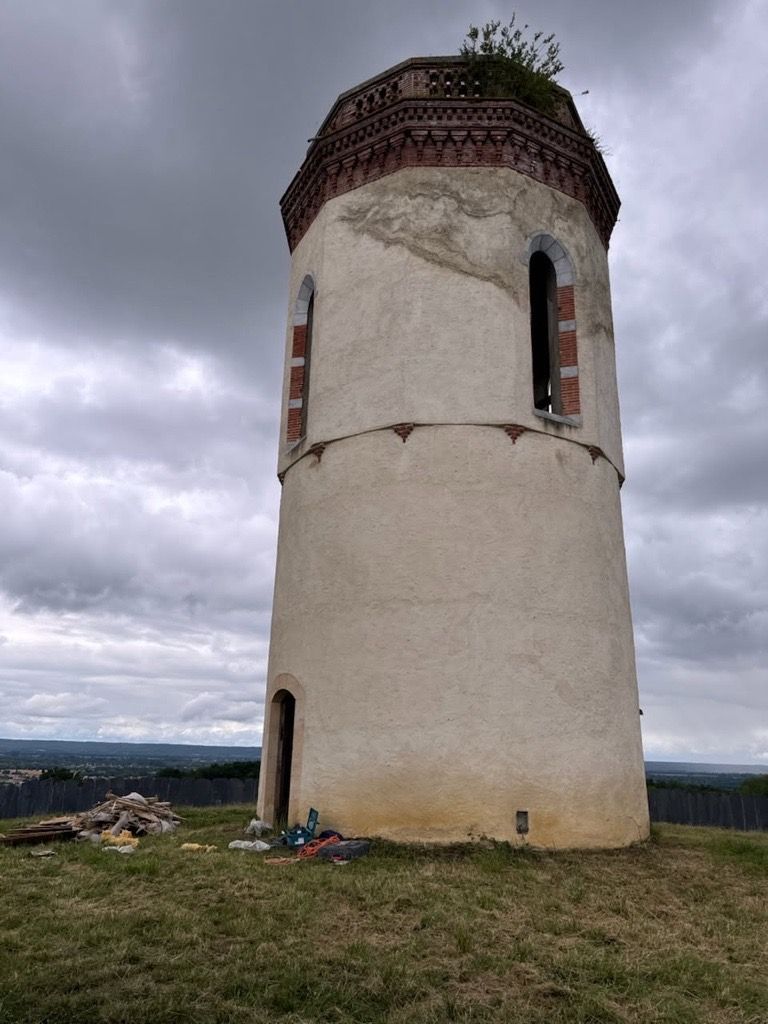 Une tour cylindrique en briques et en pierres, patinée par le temps, se dresse sur une colline herbeuse sous un ciel nuageux.