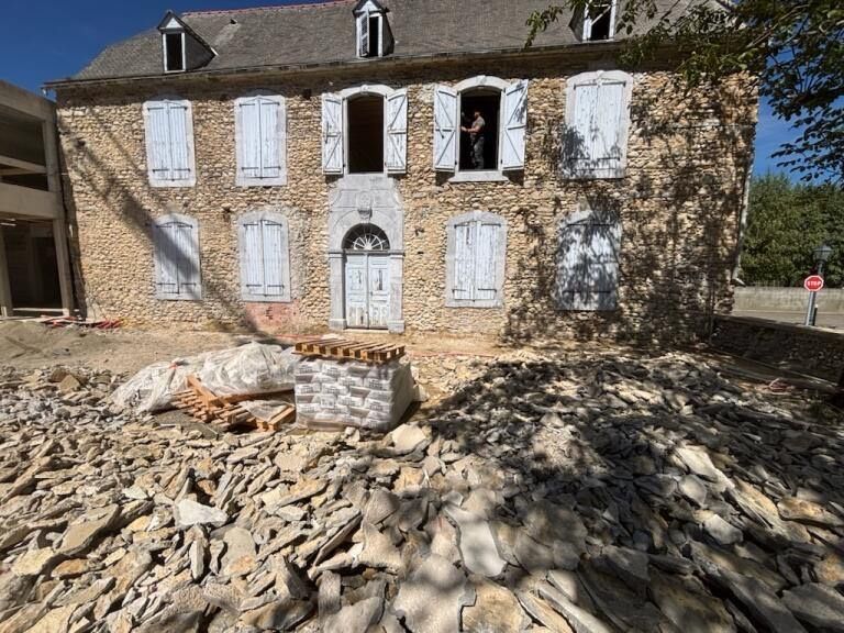 Une maison en pierre aux volets bleus en cours de rénovation, avec des débris de pierres éparses au premier plan.