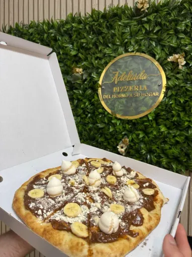 Pizza en caja, con crema de chocolate, rodajas de plátano y nata, delante de una pared verde con un letrero dorado.