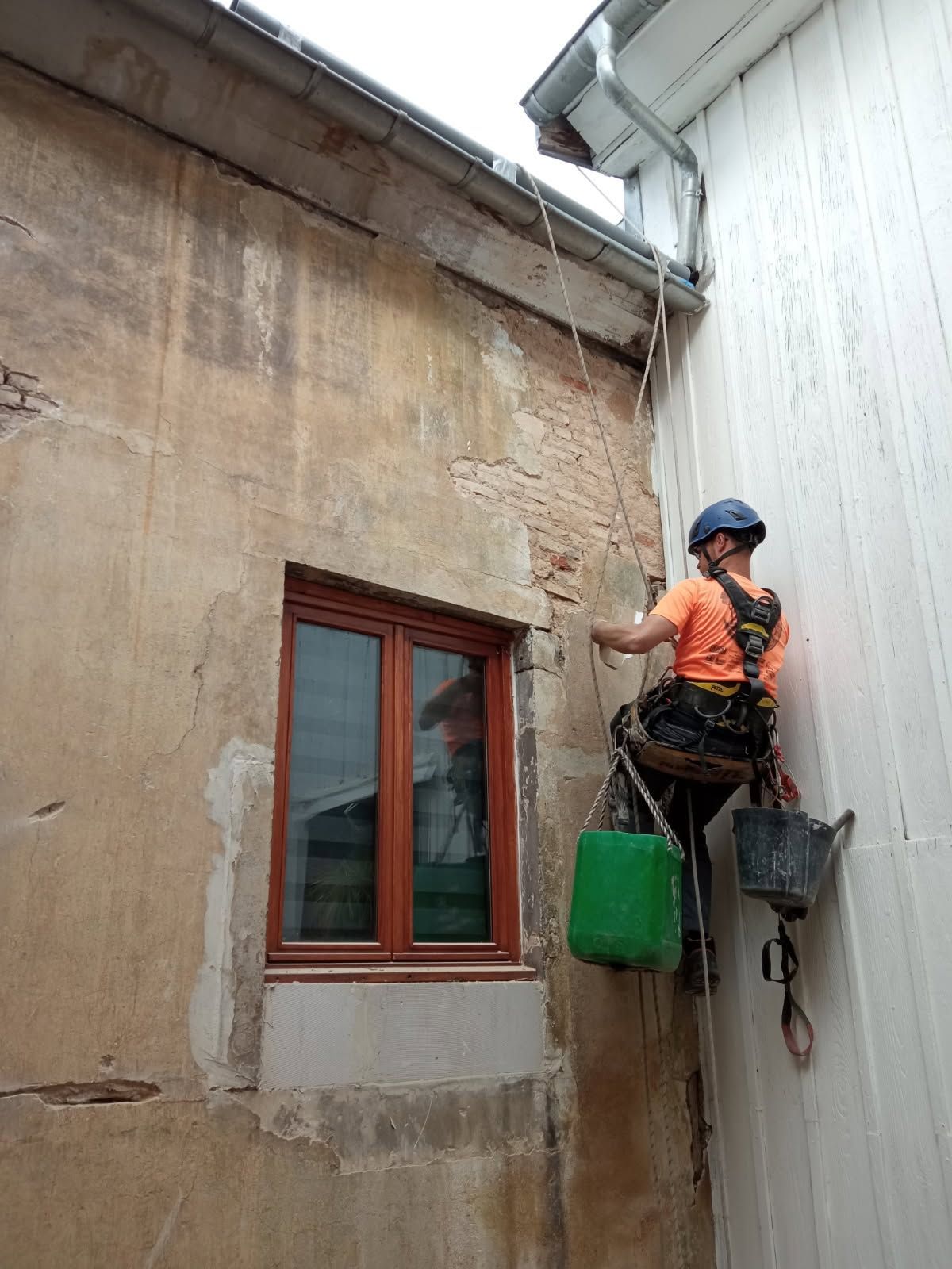 Un ouvrier, sur une plateforme suspendue, nettoie le mur d'un bâtiment à côté d'une fenêtre et d'une gouttière.