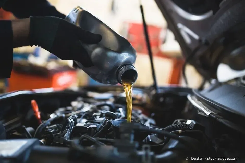 Ein Mechaniker gießt Motoröl aus einer schwarzen Flasche in den offenen Motorraum eines Autos.