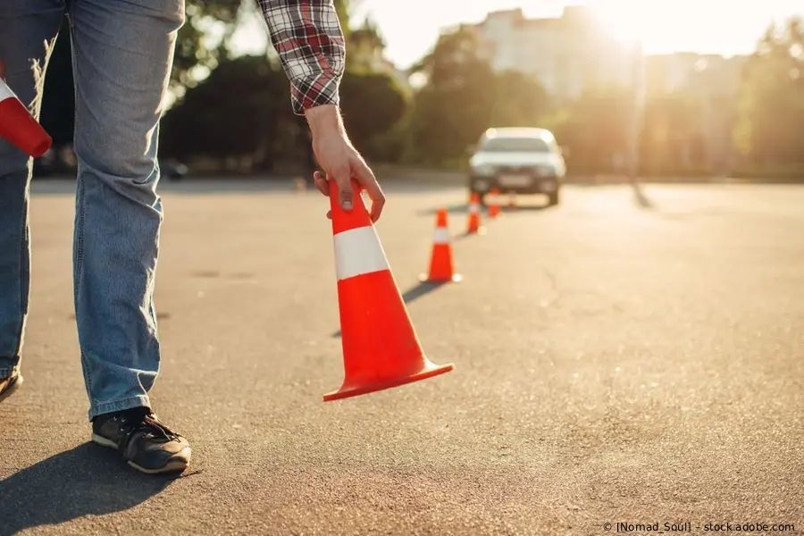 Eine Person stellt einen orangefarbenen Verkehrskegel auf eine sonnenbeschienene Straße; in der Ferne sind weitere Kegel.